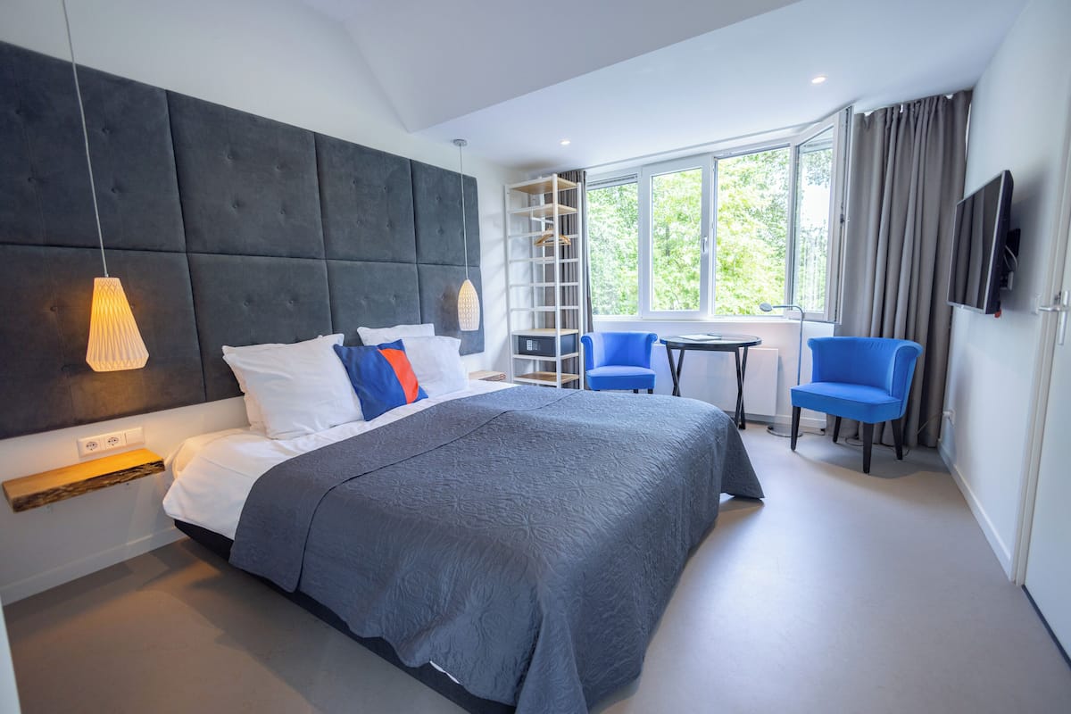 A modern hotel room with a large bed, a desk, two chairs, a TV, and a window with a view of greenery.