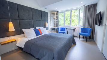 A modern hotel room with a large bed, a desk, two chairs, a TV, and a window with a view of greenery.