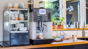 Een koffiemachine met een digitaal scherm, een glazen vitrine met keramische kopjes en een houten aanrecht met een potplant.