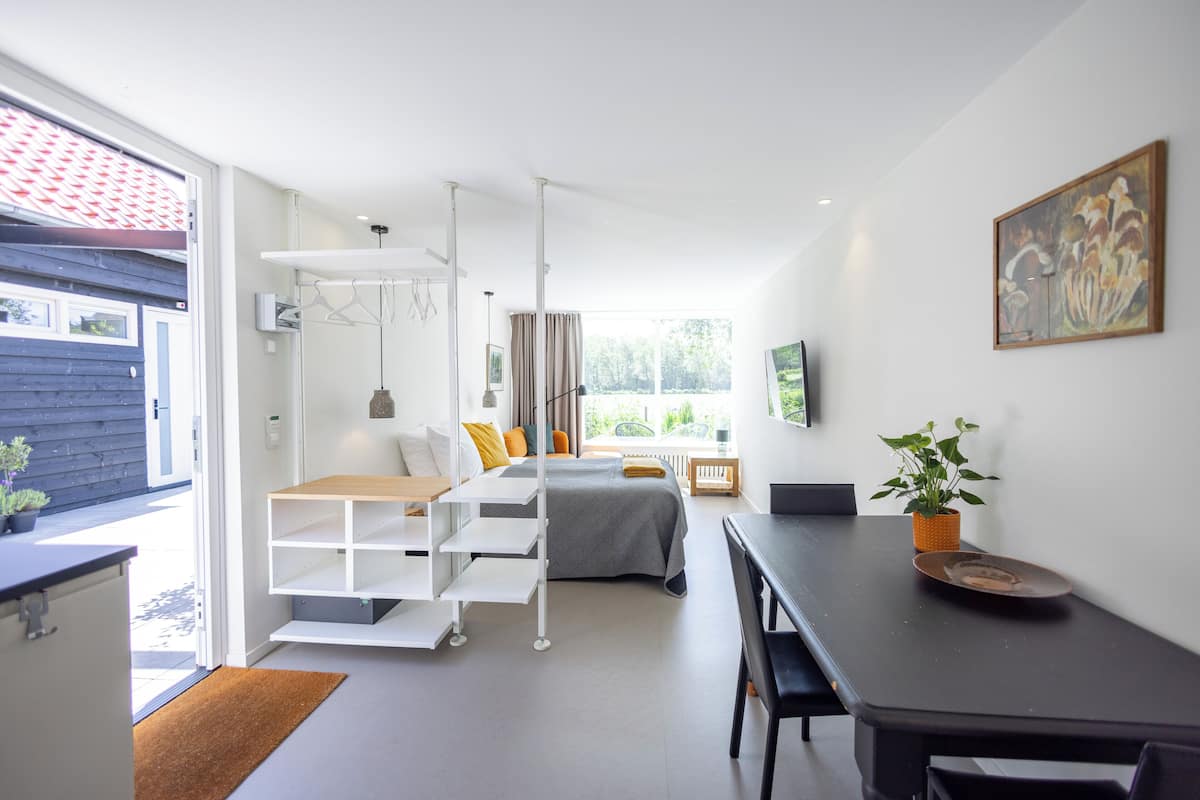 A modern bedroom with a bed, a desk, and a dining table. There is a window with curtains, a plant, and a painting on the wall.