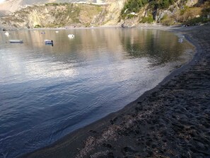 A beach with calm water, boats, and a rocky hillside in the background.