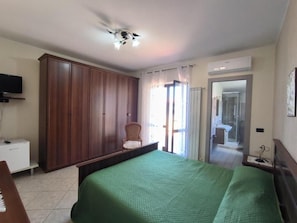 A bedroom with a bed, a wardrobe, a chair, a television, and a bathroom visible through a glass door.