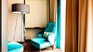 Una habitación con un sillón azul turquesa, una lámpara de pie y un gran ventanal con cortinas.