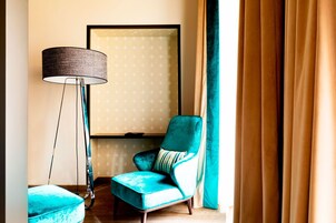 Una habitación con un sillón azul turquesa, una lámpara de pie y un gran ventanal con cortinas.