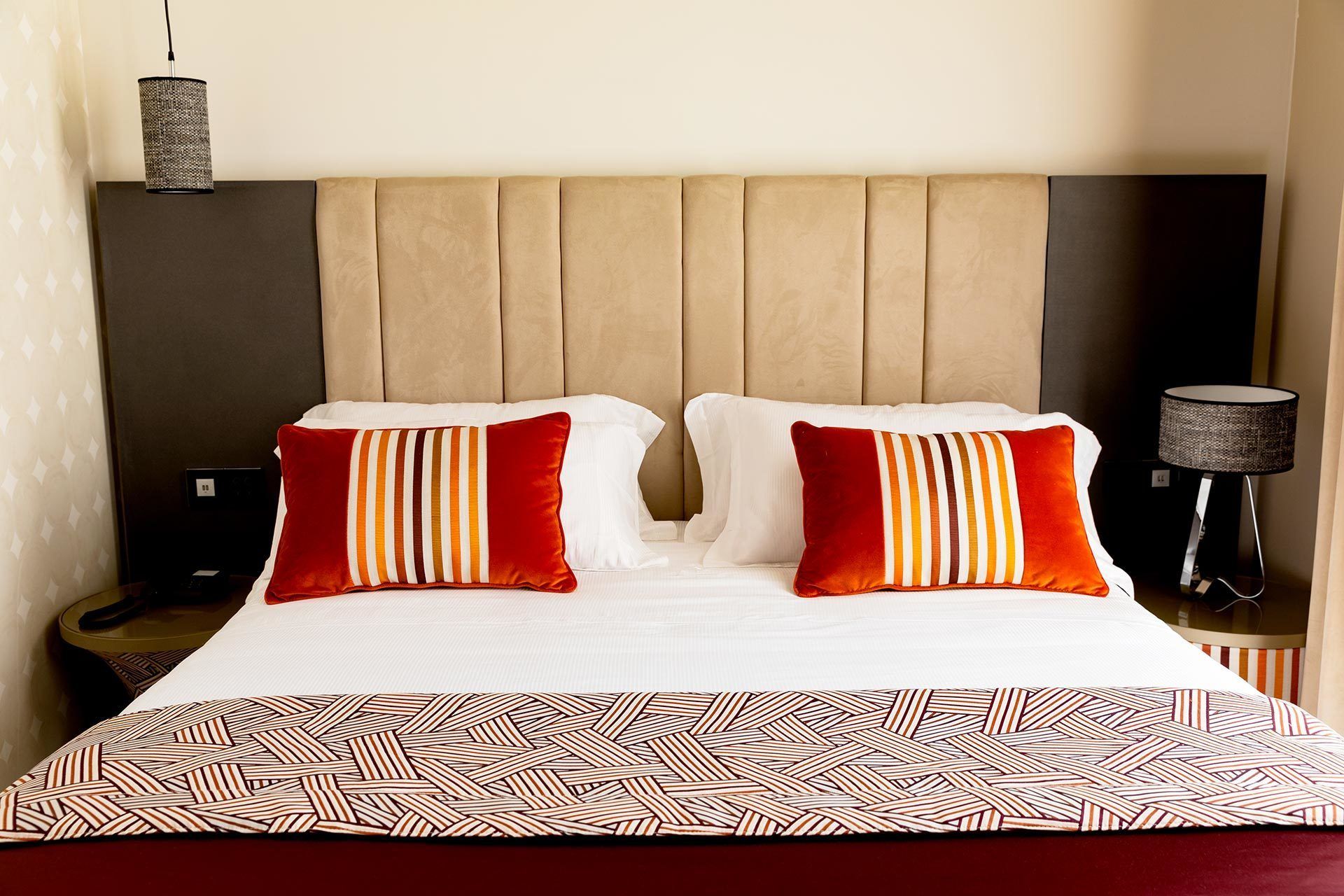 A hotel room with a large bed, two pillows with striped patterns, a patterned throw, a lamp, and a speaker on the nightstand.