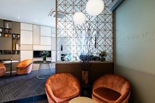 Un interior moderno con un diseño geométrico en la pared, una zona de estar con dos sillones acolchados, una mesa redonda y un jarrón decorativo con ramas.