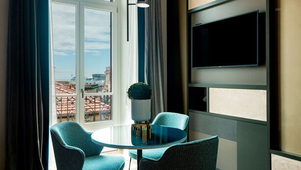 Una habitación moderna con una mesa de vidrio y dos sillas, un ventanal con cortinas y un televisor montado en la pared.