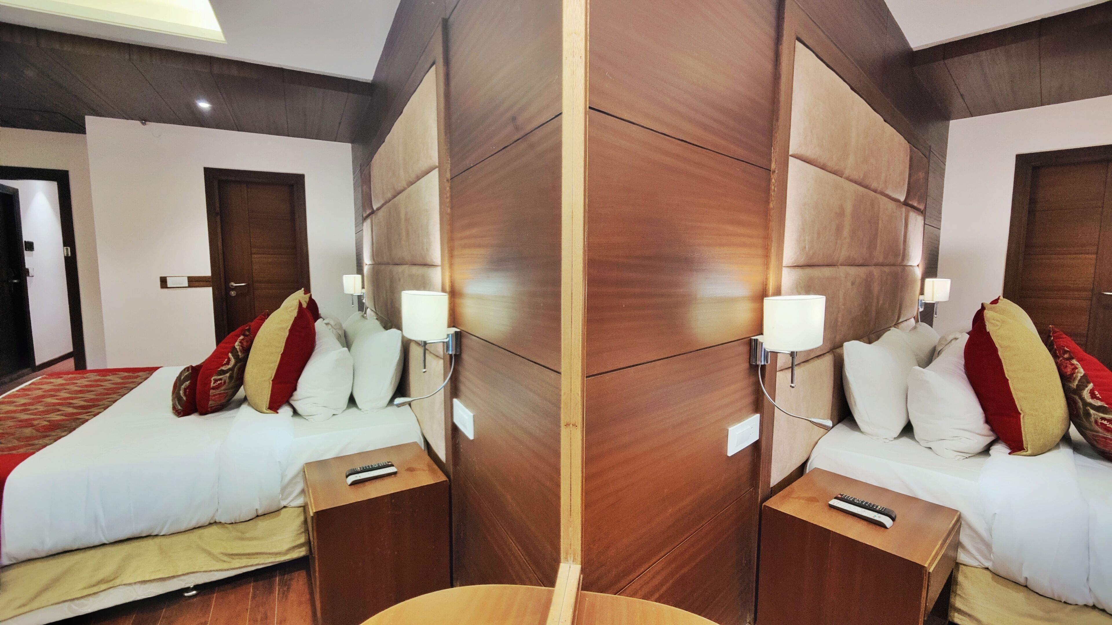 Een hotelkamer met spiegel, een houten hoofdbord, een bed met wit beddengoed en rode en gouden kussens, bedlampjes en een afstandsbediening op het nachtkastje.