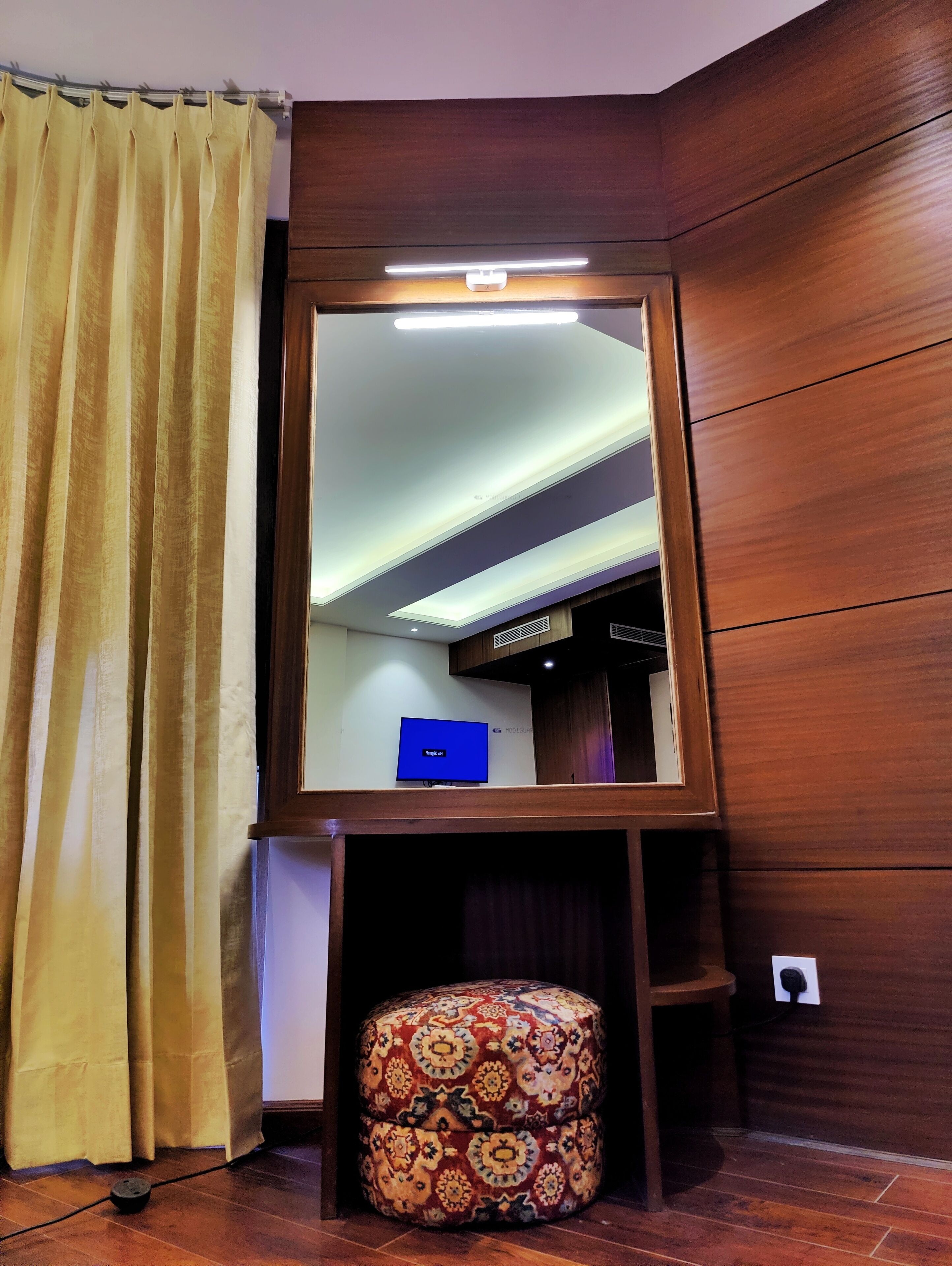 A room with a large mirror, wooden walls, a patterned ottoman, and a television on a stand.