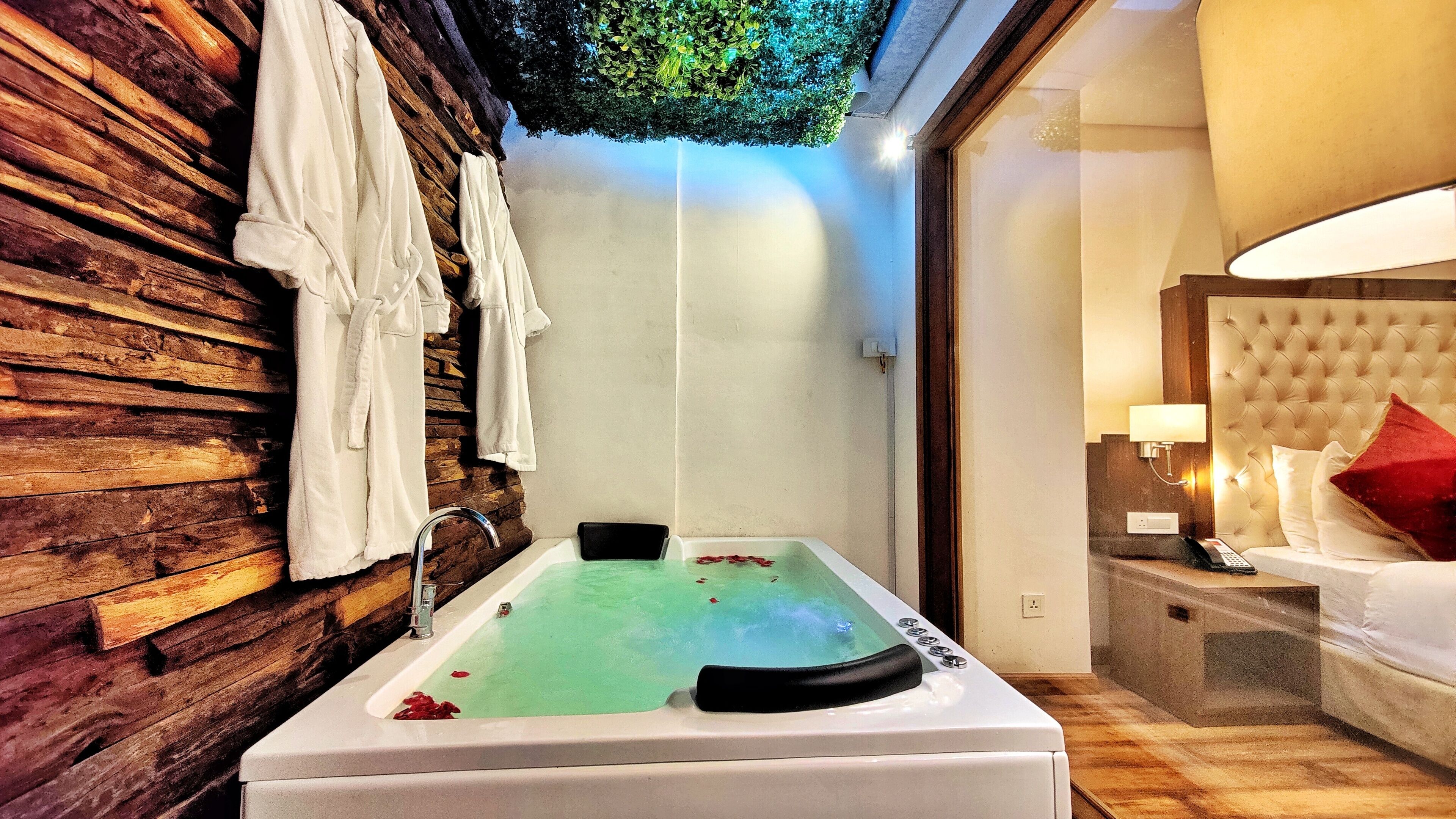 A modern bathroom with a wooden wall, a bathtub with rose petals, a mirror, and a bedside table with a lamp.