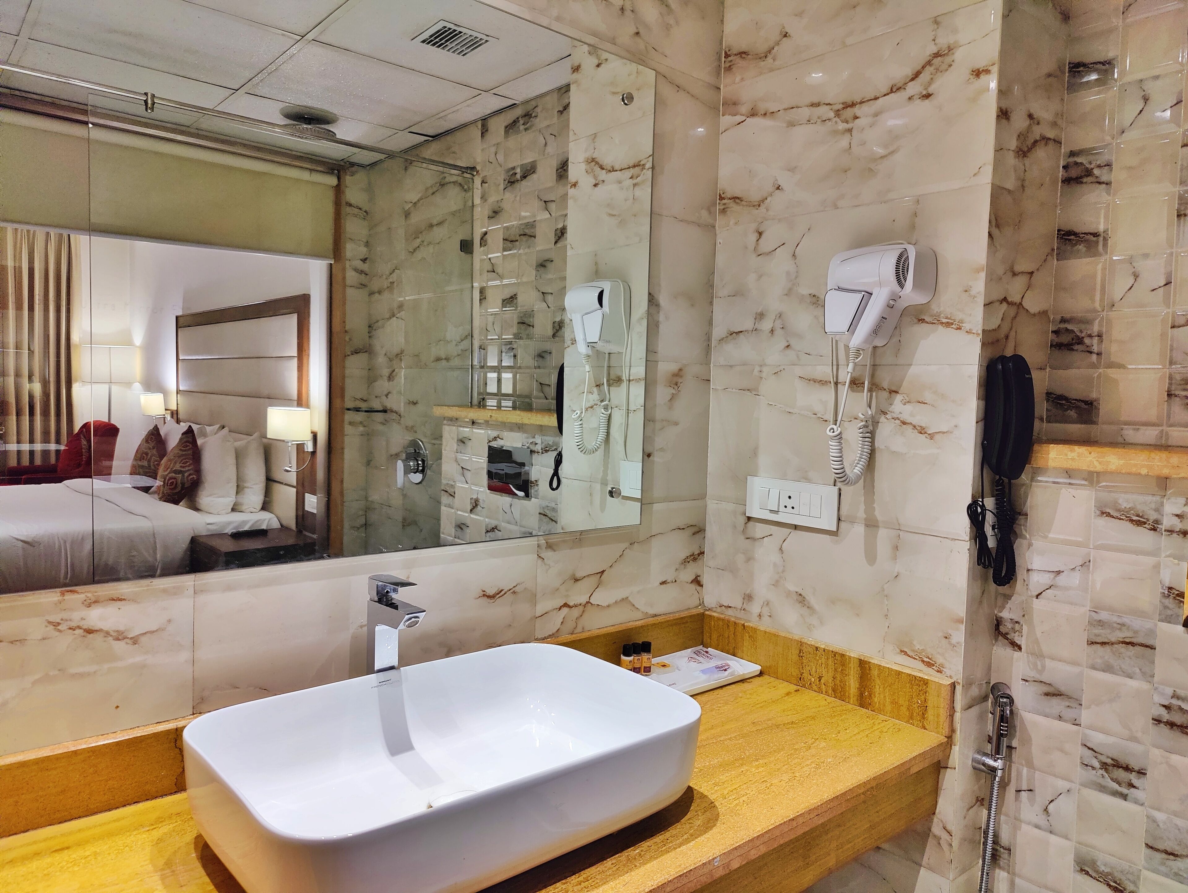 A hotel bathroom with a marble wall, a large mirror, a white sink, a hairdryer, and a telephone.