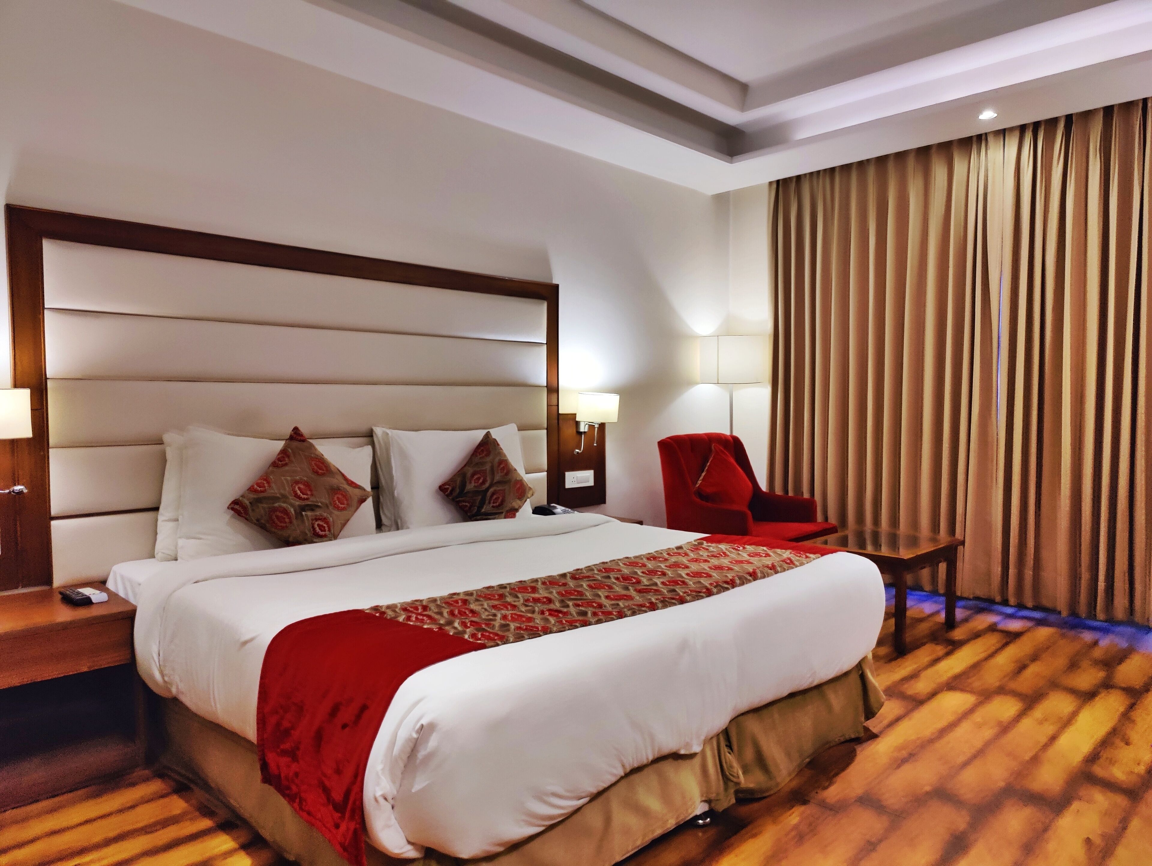 A hotel room with a large bed, a red armchair, a wooden floor, and a curtain-covered window.