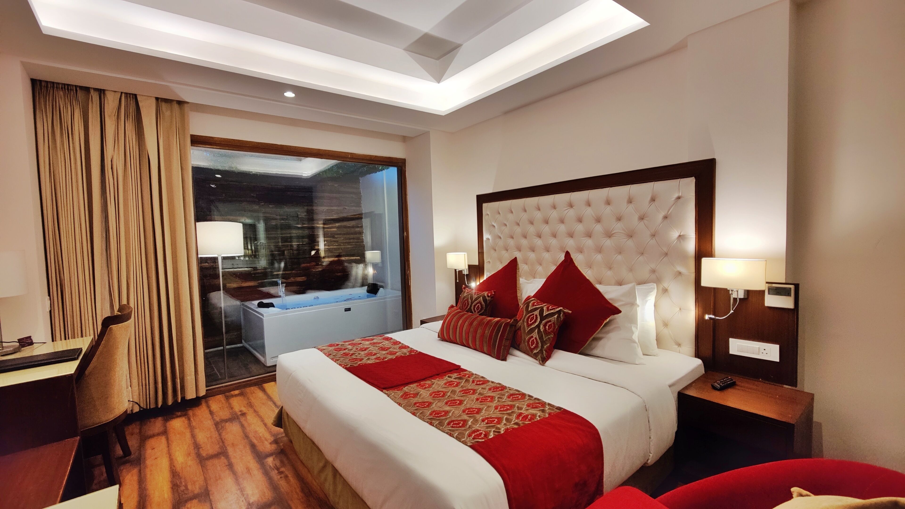 Une chambre d’hôtel avec un grand lit, un bureau, une chaise, une lampe de bureau, une table de chevet, une commode, une lampe fixée au mur et une fenêtre avec des rideaux.