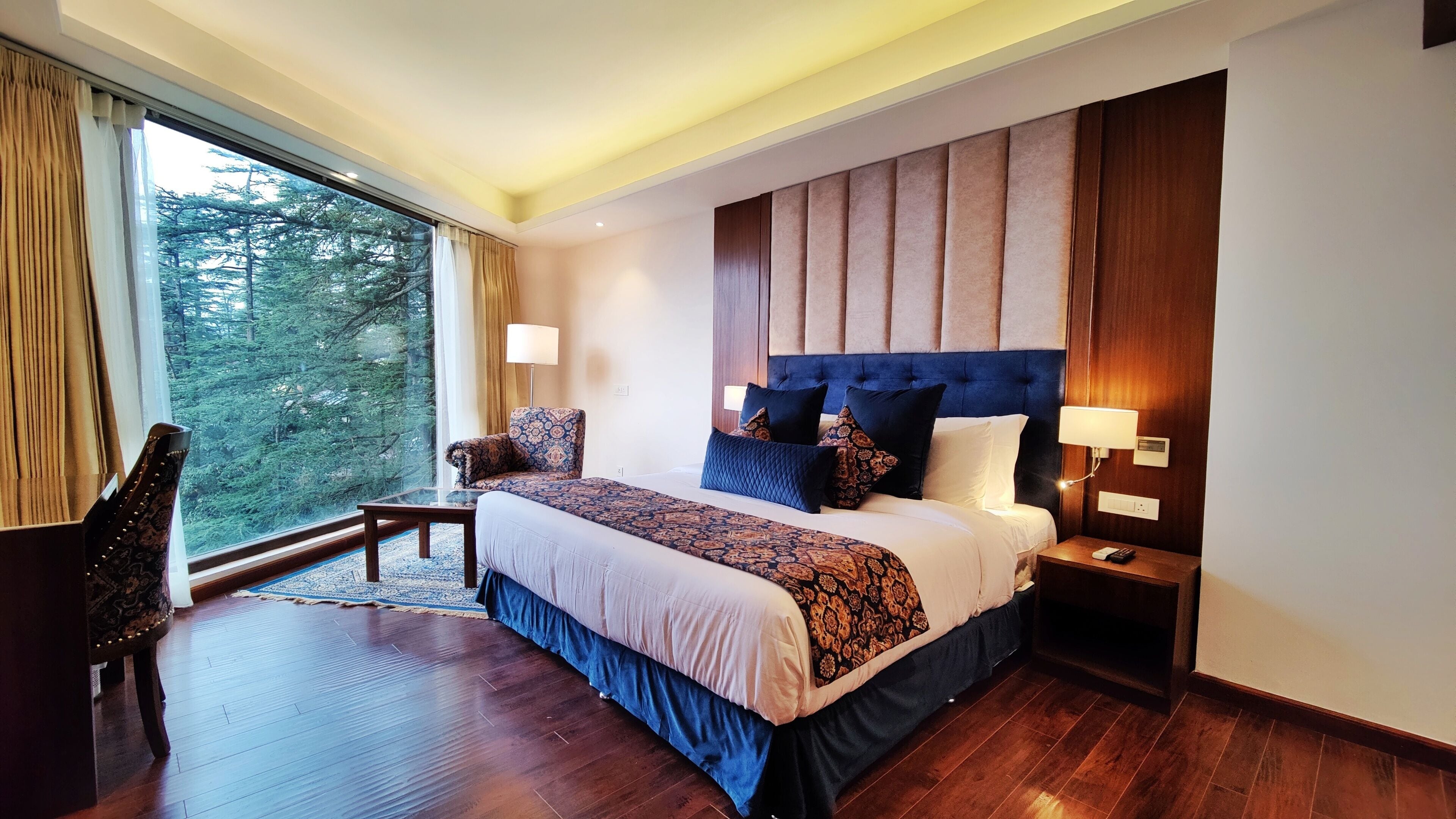 A hotel room with a large bed, a nightstand, a chair, and a view of trees through a large window.