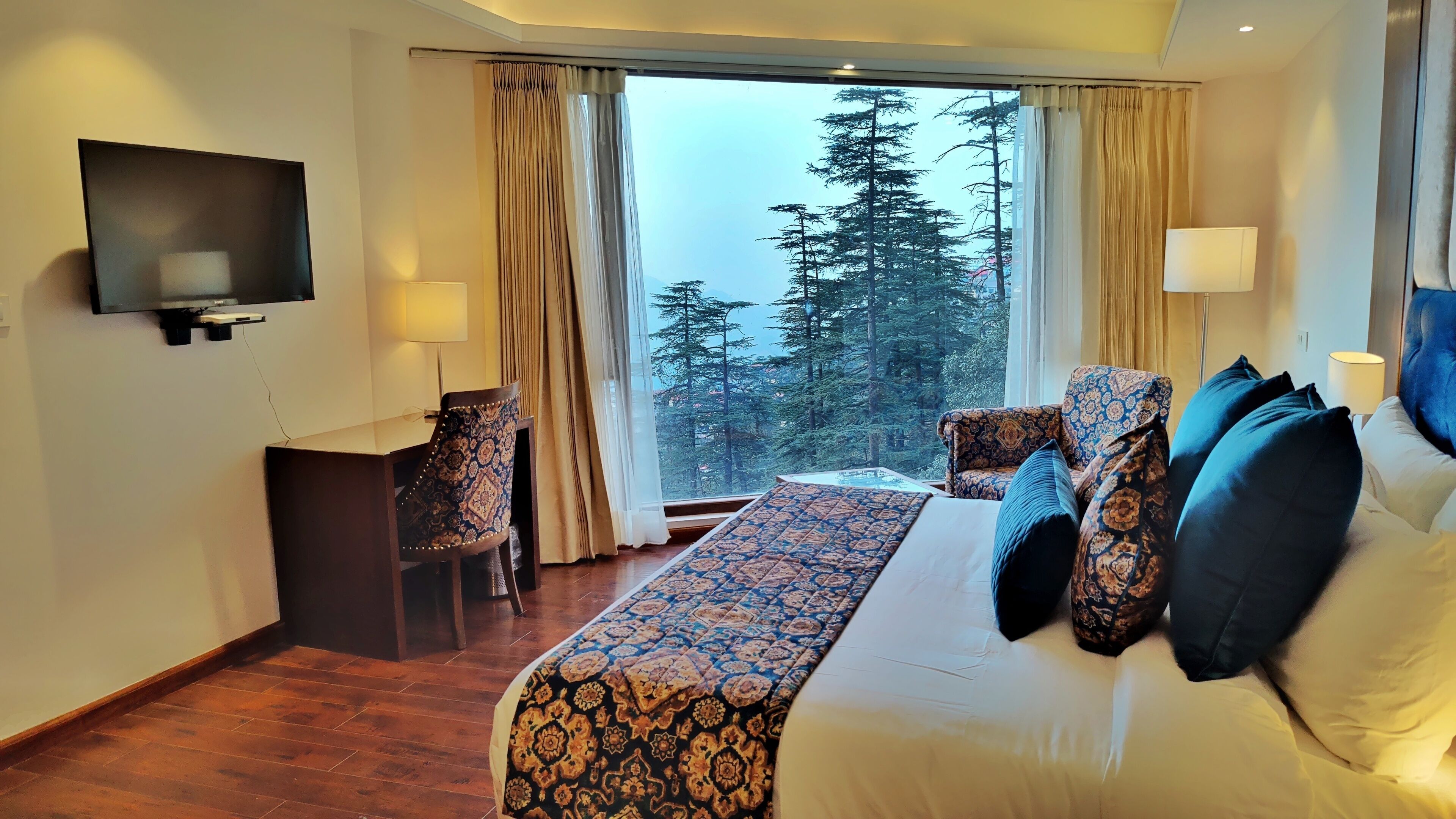 A hotel room with a large bed, a desk with a chair, a TV mounted on the wall, and a window with curtains.