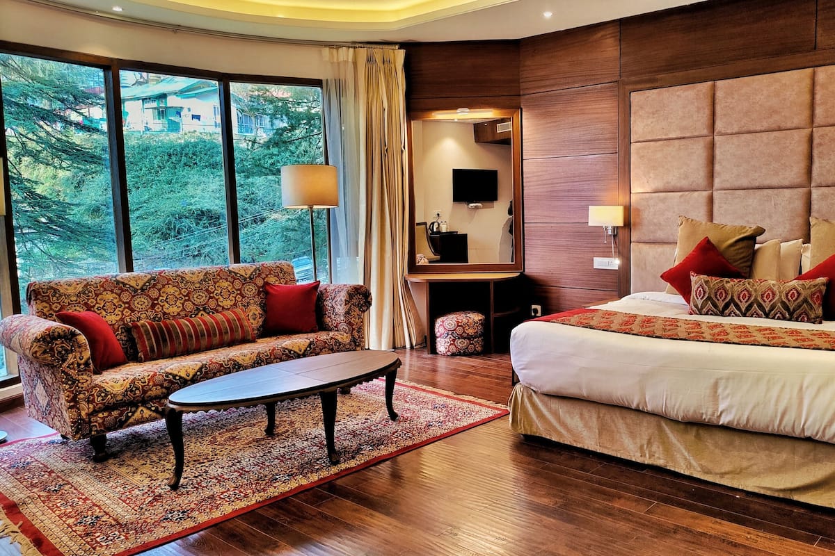 A hotel room with a sofa, a bed, a coffee table, a rug, and a large window with a view of greenery.