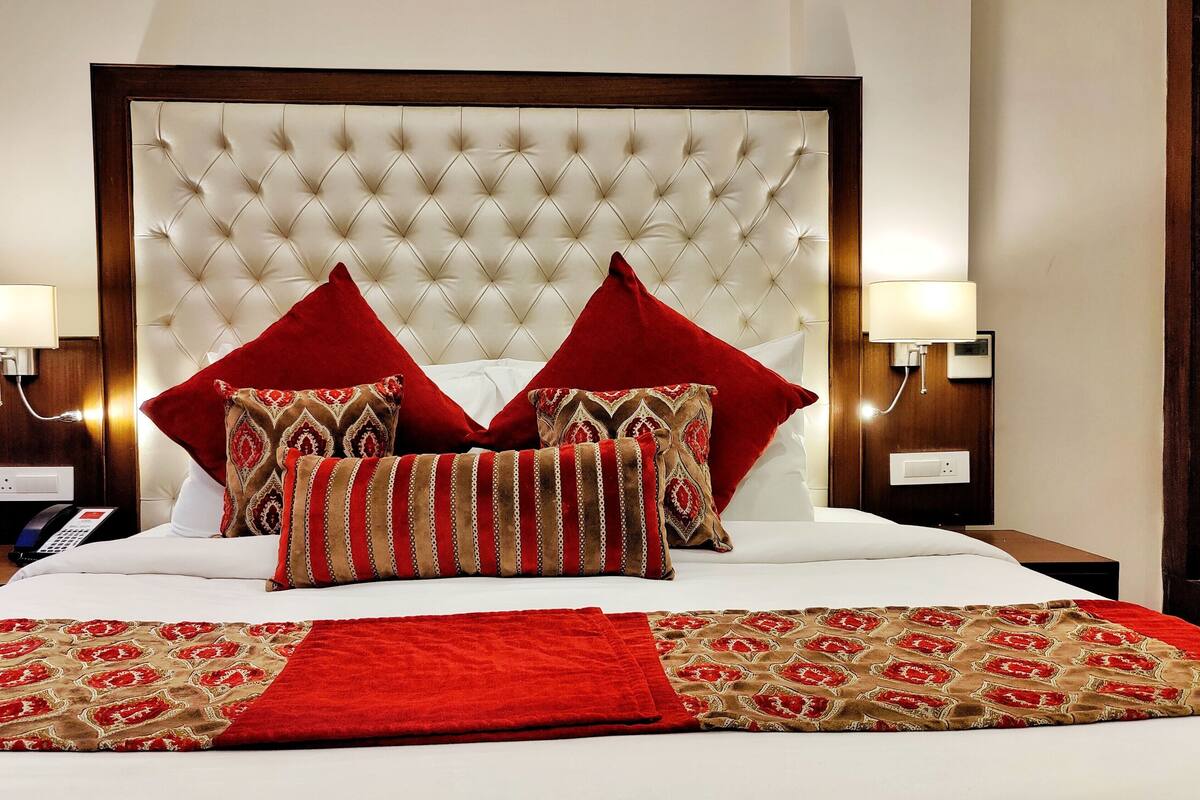 A hotel room with a tufted headboard, two bedside lamps, a telephone, and a bed with red and patterned bedding.