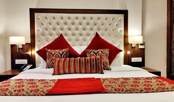 Una habitación de hotel con cabecera capitonada, dos lámparas de noche, un teléfono y una cama con ropa de cama roja y estampada.