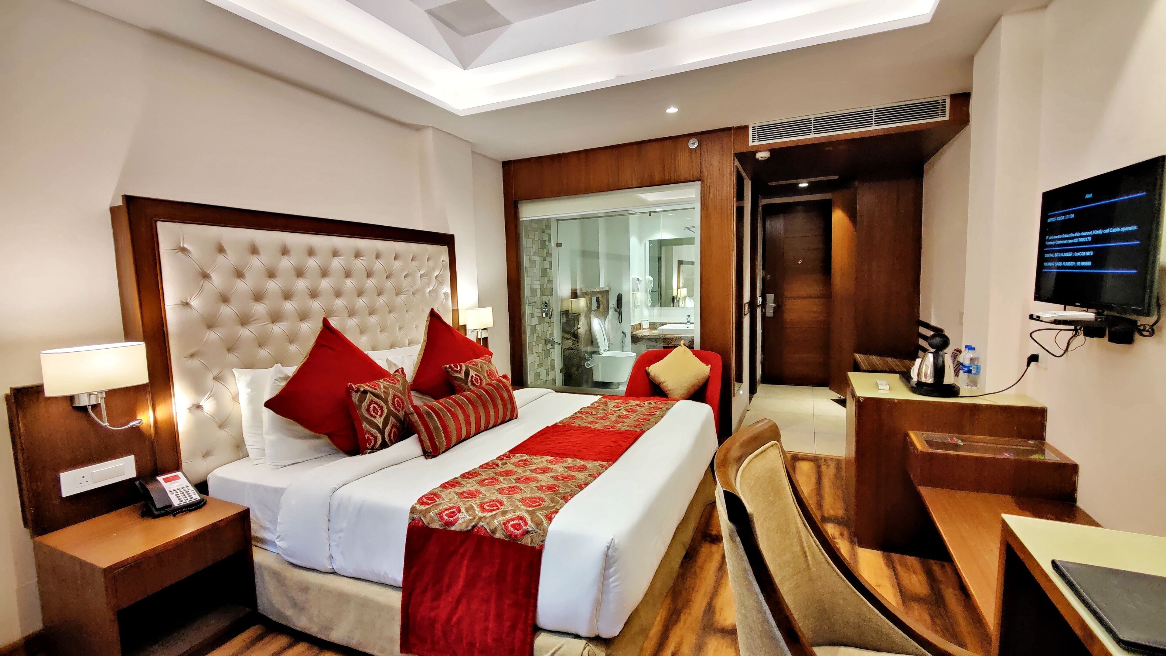 A hotel room with a large bed, a desk, a TV, and a bathroom visible through a glass door.