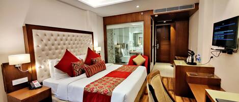 A hotel room with a large bed, a desk, a TV, and a bathroom visible through a glass door.