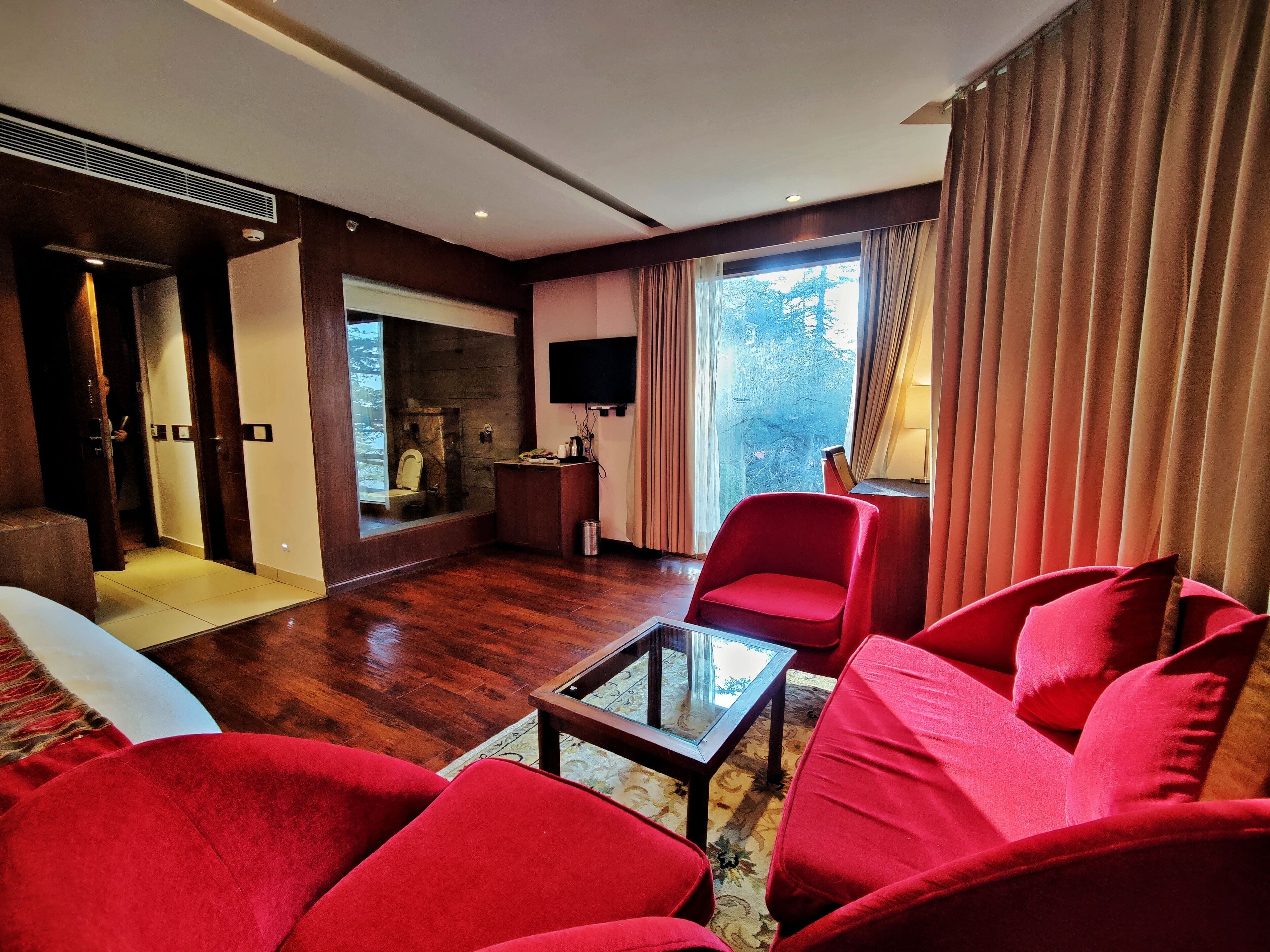 Una sala de estar moderna con un sofá rojo, una mesa de centro de vidrio y una chimenea. Hay un televisor montado en la pared y una ventana con cortinas.