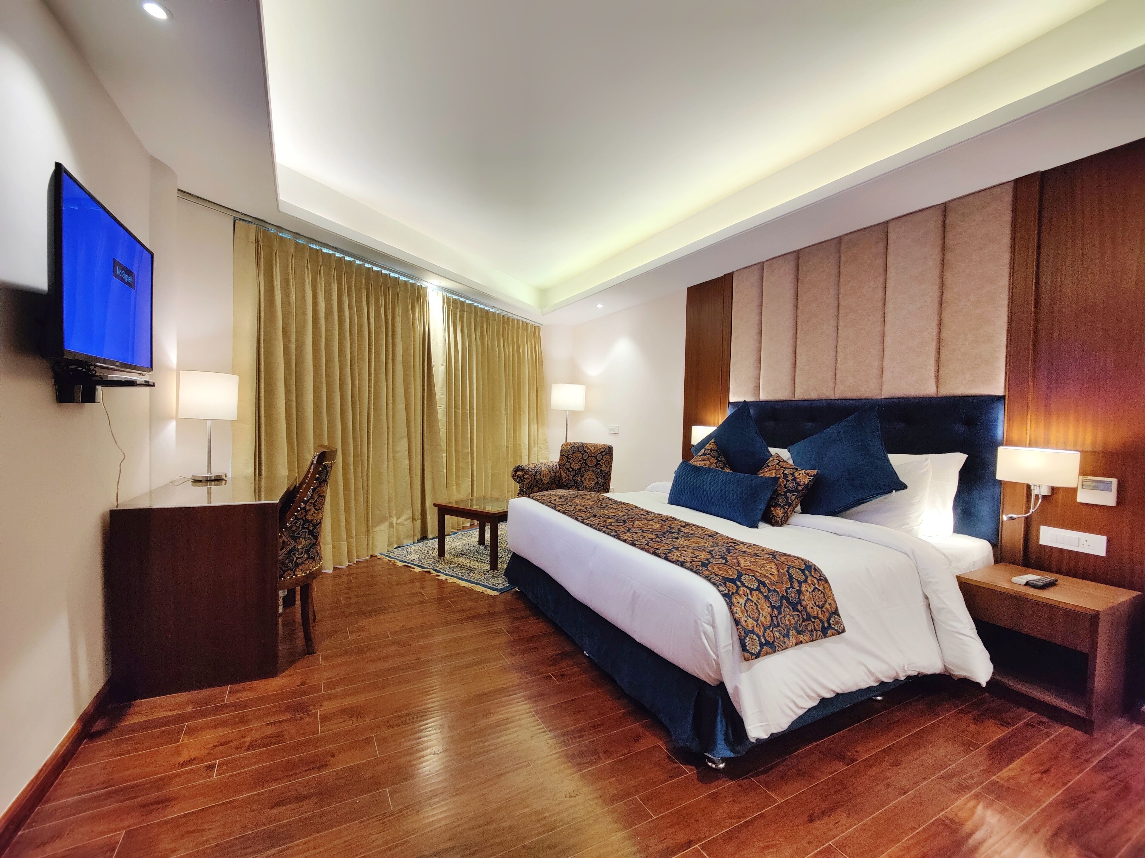 A well-lit hotel room with a large bed, a television, a desk, and a chair.