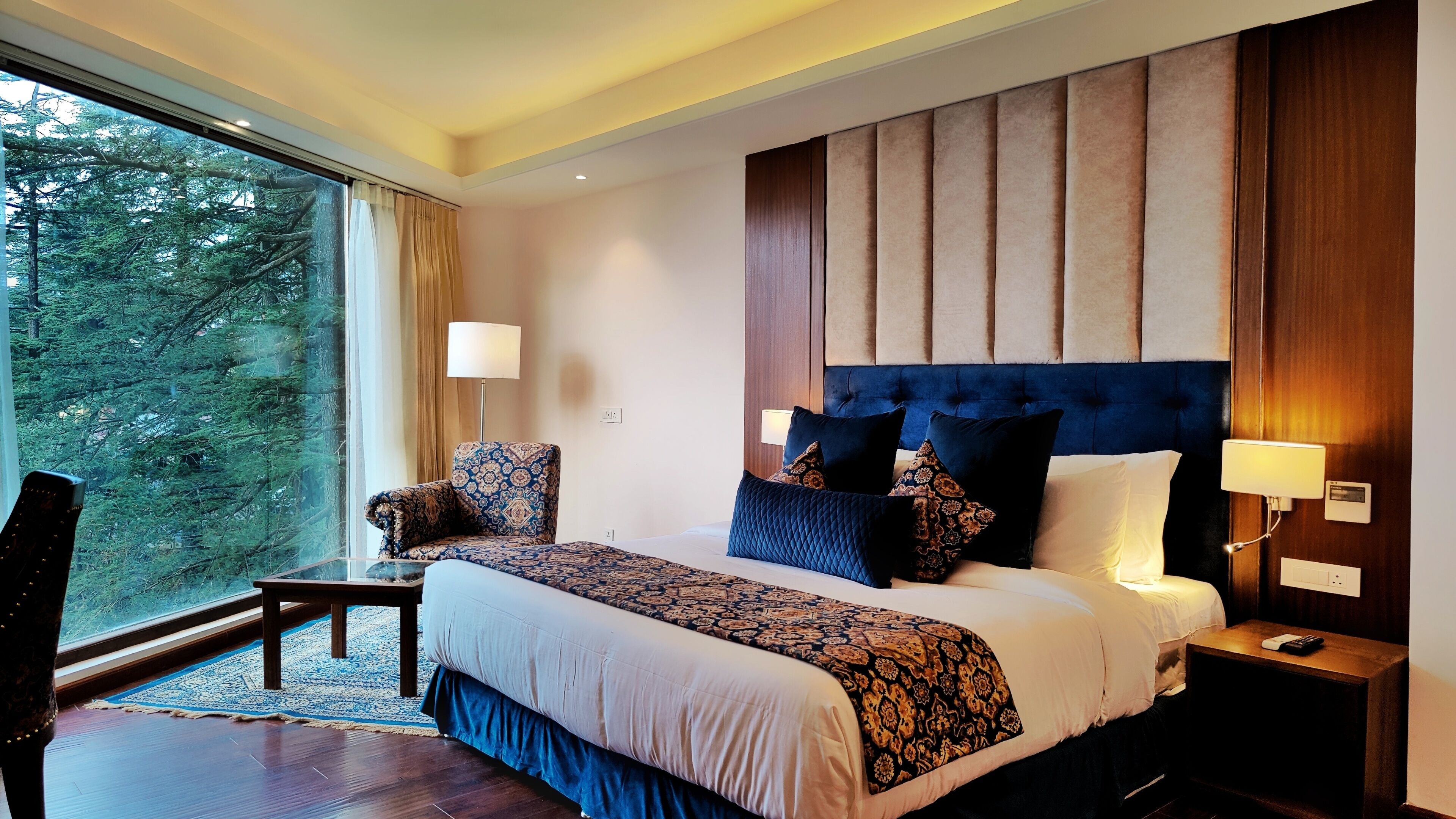 A hotel room with a large bed, a chair, a small table, and a view of trees through the window.