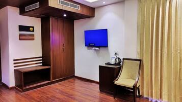 A hotel room with a wooden bench, a TV mounted on the wall, a small table with a kettle, and a chair.