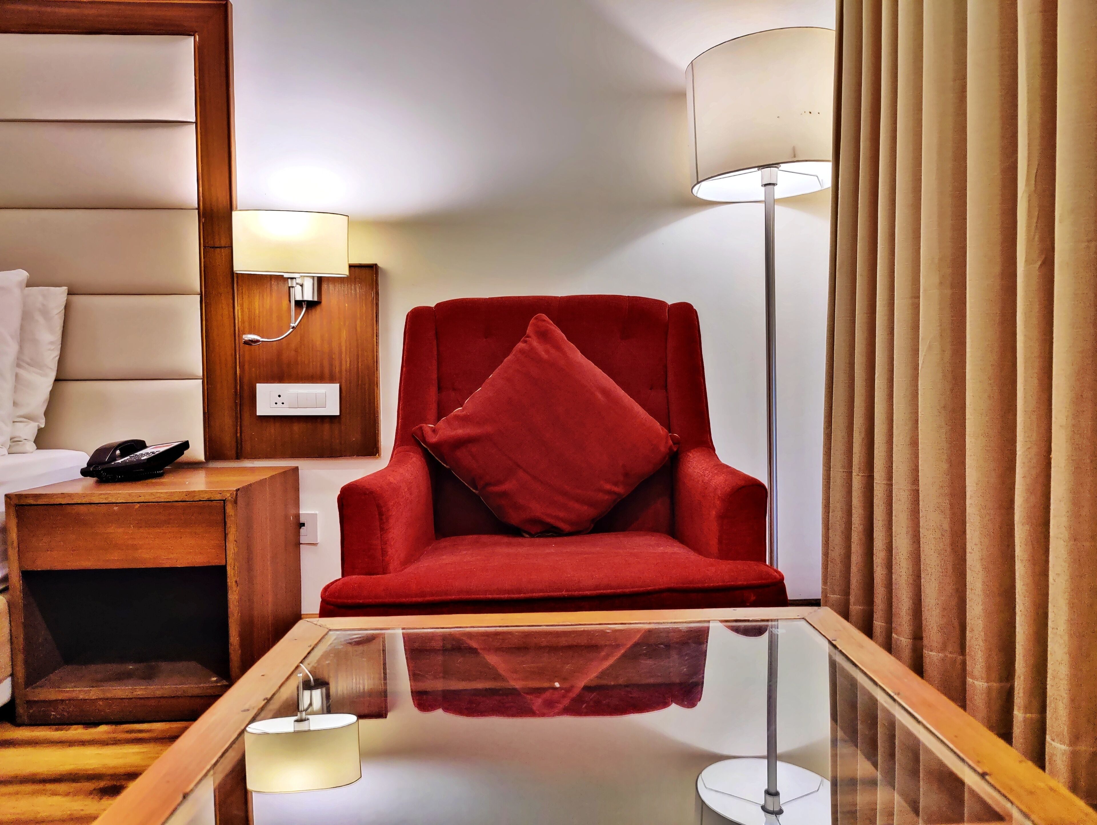 A hotel room with a red armchair, a bedside table with a lamp, a phone, and a glass-top table with a candle.
