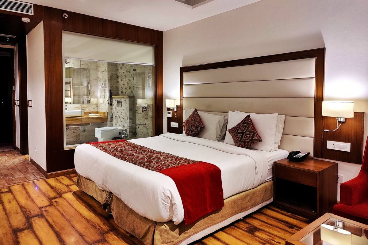 A hotel room with a large bed, a bedside table with a lamp, a red chair, and a bathroom visible through a glass door.