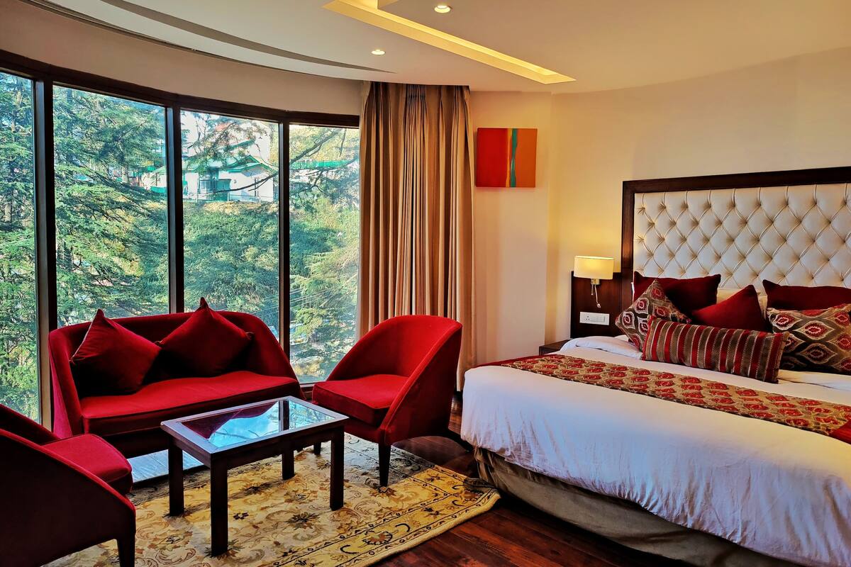 A hotel room with a large window, a bed with red and patterned pillows, a red sofa, and a small table.