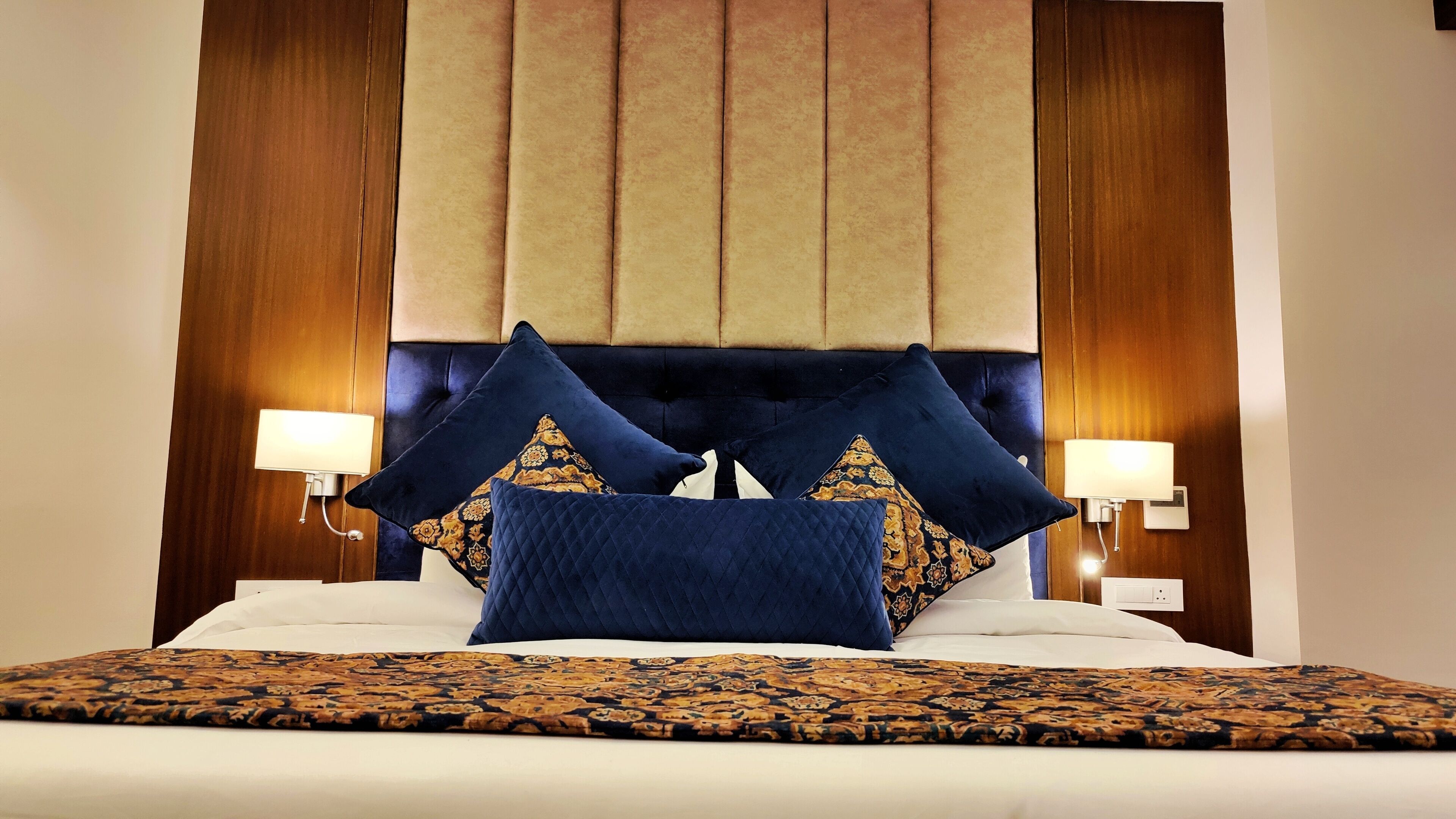 A bed with a dark blue headboard, two decorative pillows, a patterned bedspread, and two bedside lamps.