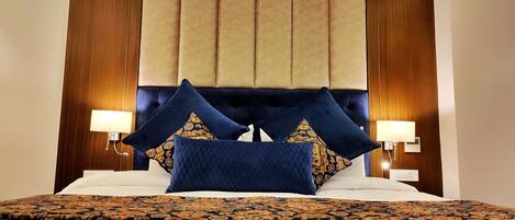 A bed with a dark blue headboard, two decorative pillows, a patterned bedspread, and two bedside lamps.