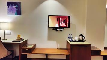A hotel room with a flat-screen TV mounted on the wall, a desk with a lamp, a bench, and a small table with a coffee maker and water bottles.
