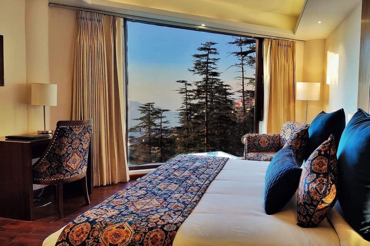 A hotel room with a large bed, a desk, and a chair. There is a view of trees and mountains through the window.