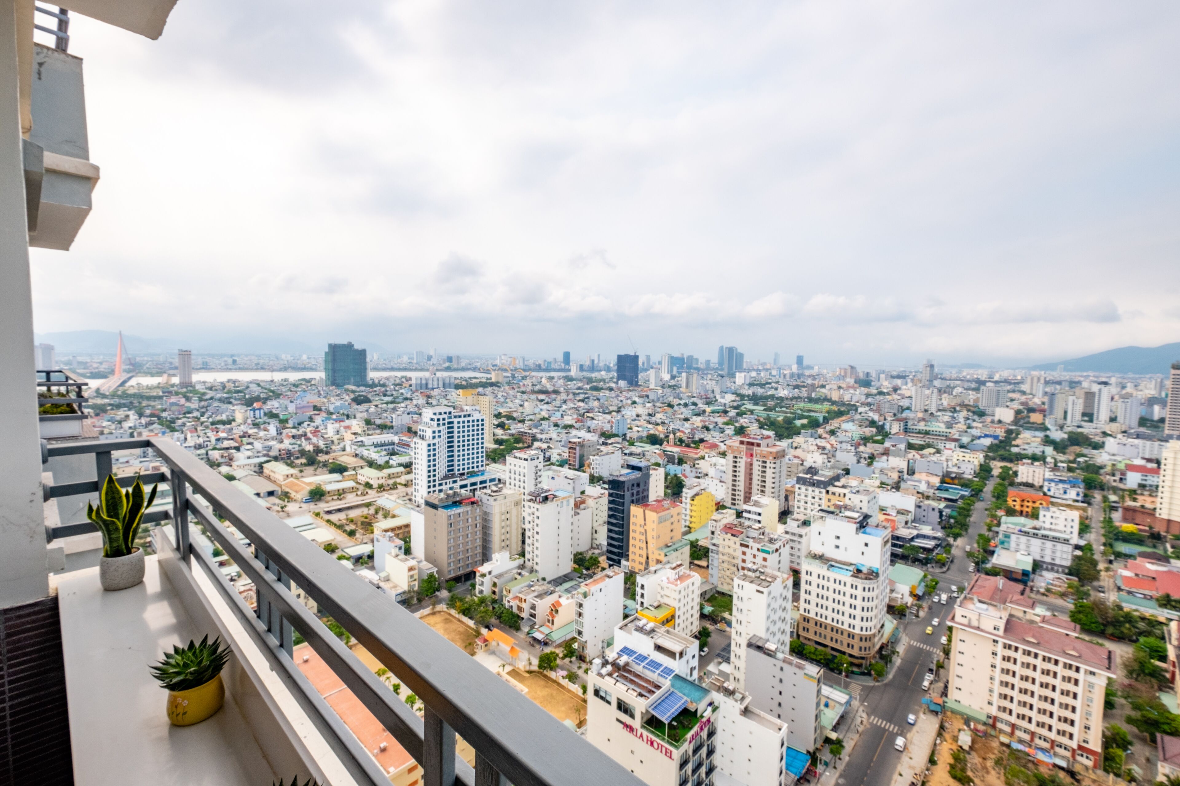 A cityscape view from a balcony with potted plants, featuring a mix of residential and commercial buildings, a prominent bridge, and distant mountains under a cloudy sky.