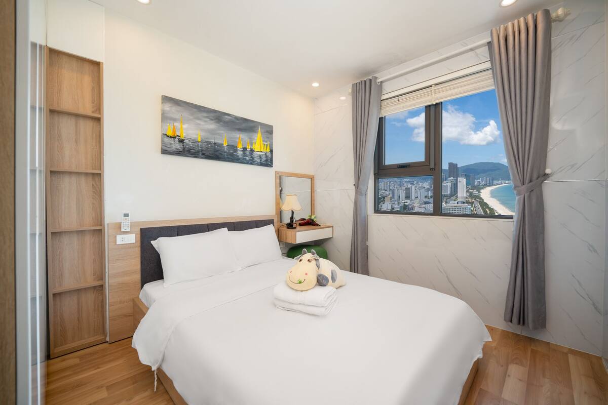 A hotel room with a bed, a painting of sailboats, a city view, and a window with curtains.