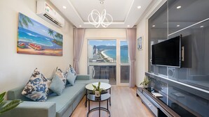 A modern living room with a sofa, a coffee table, and a TV. There is a painting of a beach scene on the wall, and a view of a cityscape through large windows. - Apartment SeaView at Muong Thanh Residence (Da Nang)