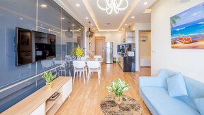 A modern living room with a flat-screen TV, a blue sofa, and a dining area with a table and chairs. - Apartment SeaView at Muong Thanh Residence (Da Nang)