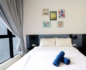 Habitación de hotel con cama, ropa de cama blanca y dos toallas azules enrolladas. Hay obras de arte en la pared con motivos náuticos.