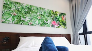 Una cama bien hecha con sábanas blancas y dos toallas azules enrolladas, un cabecero de madera, un cuadro colgado en la pared con hojas y flores tropicales, y una ventana con cortinas.