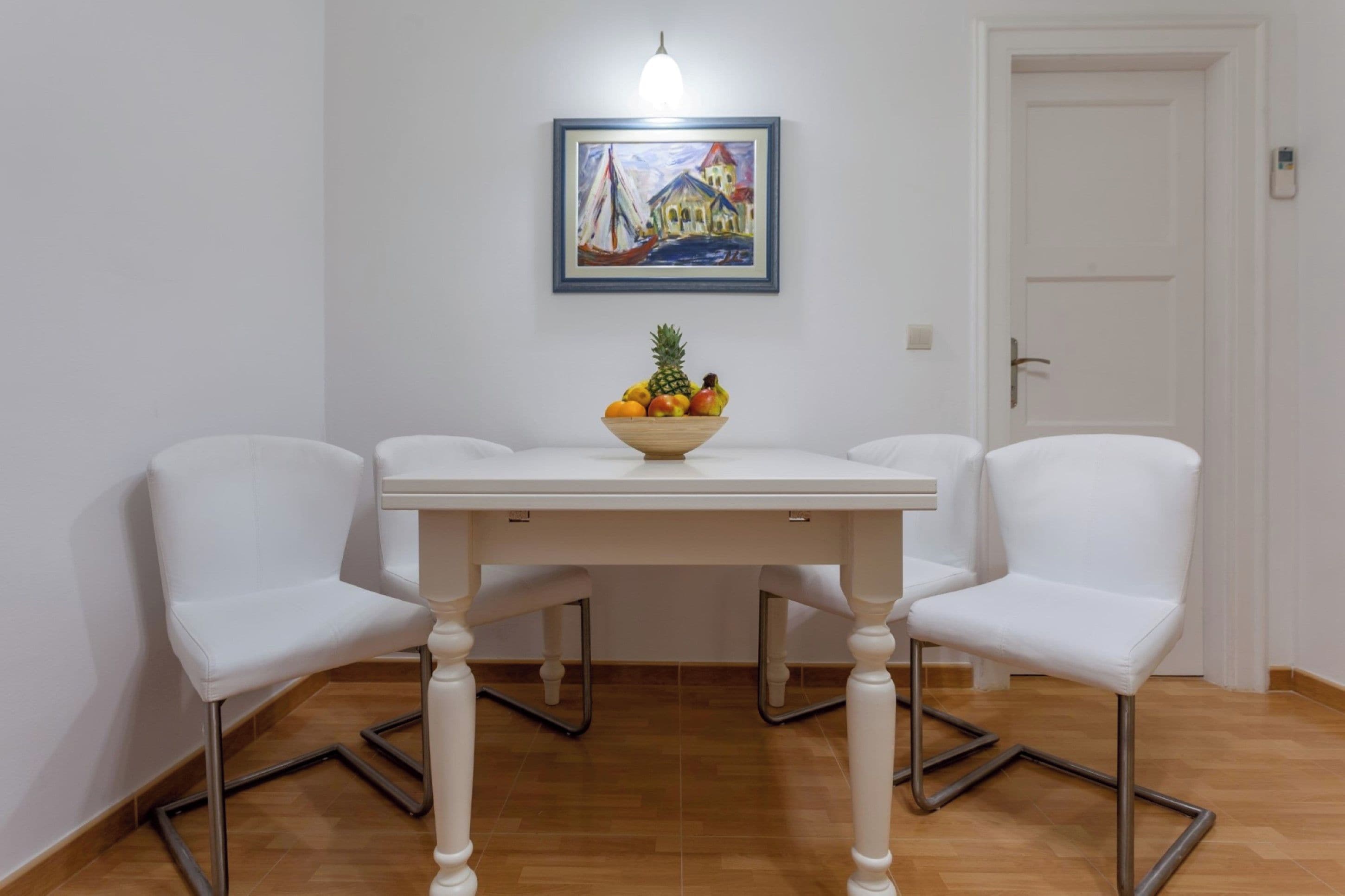 Un coin repas avec une table blanche, deux chaises blanches, un panier de fruits et un tableau accroché au mur.