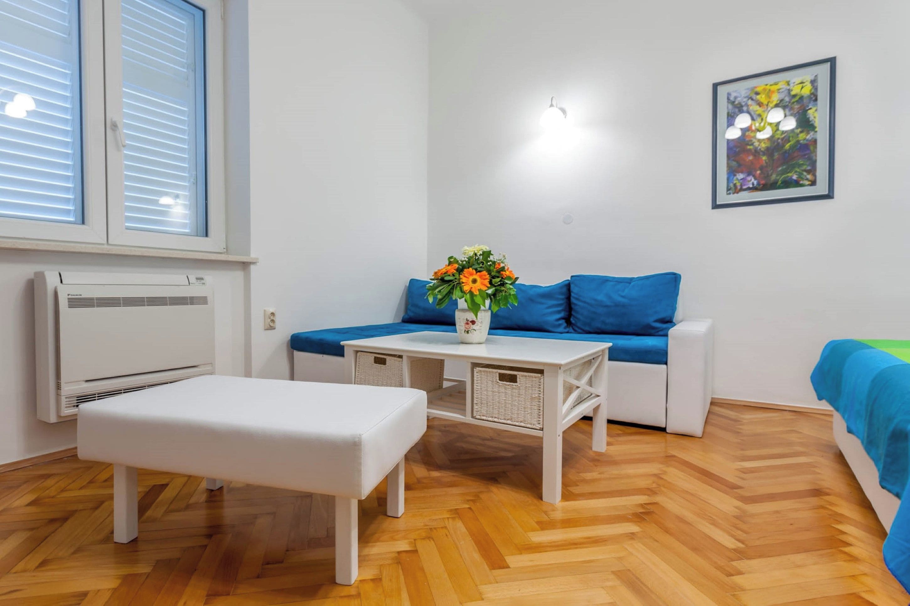 A room with a white bench, a blue sofa, a small table with a vase of flowers, and a wall-mounted light fixture.