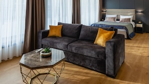 Ein modernes Hotelzimmer mit einem dunkelgrauen Sofa, einem Glastisch und einem Bett mit einer blau-grauen Tagesdecke.