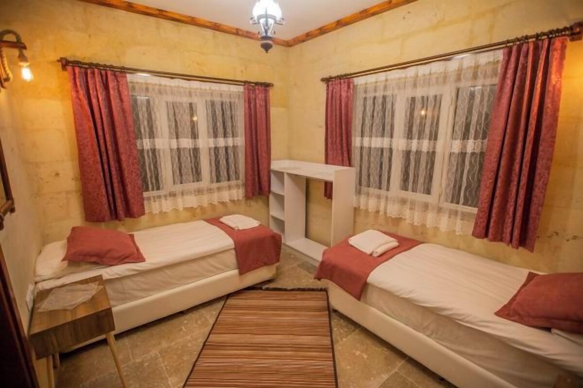A room with two beds, a small table, a shelf, and a ceiling light.