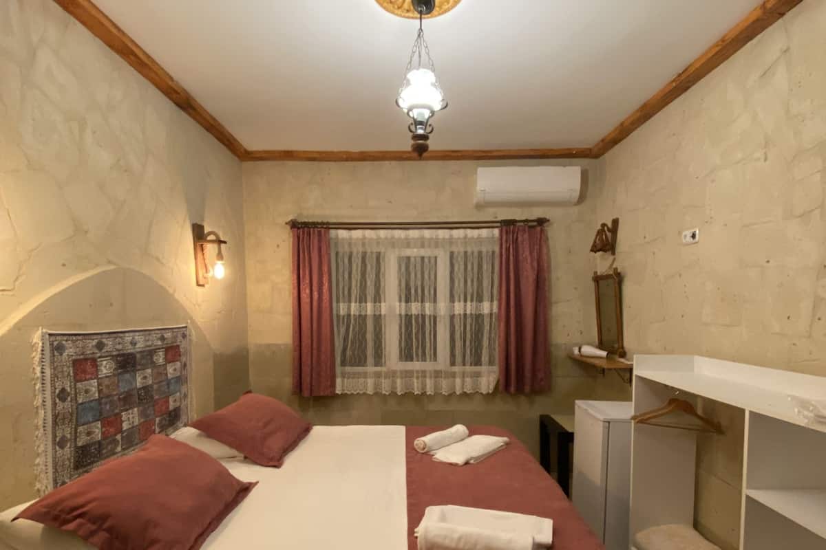 Ein Schlafzimmer mit Bett, einer Pendelleuchte, einem Fenster mit Vorhängen, einer Wandtäfelung mit Patchwork-Quilt und einem kleinen weißen Schrank.