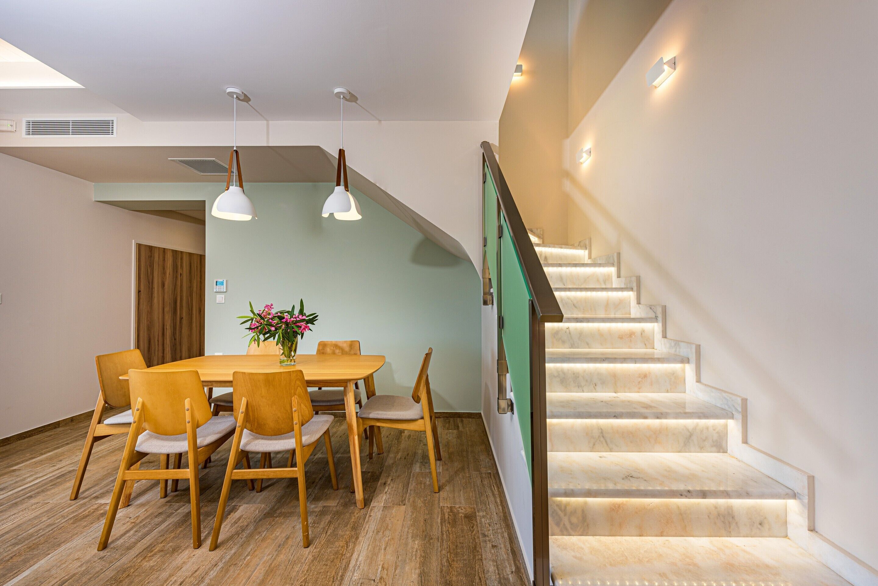 A dining area with a wooden table and chairs, a vase with flowers, and a staircase with illuminated steps.