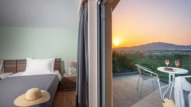Une chambre d’hôtel comprenant un lit, une table de chevet, une lampe, un balcon avec une table et des chaises, et une vue sur le coucher du soleil se reflétant sur un paysage.