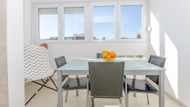 Un coin repas avec une table en verre, quatre chaises, un saladier de fruits et une fenêtre avec des stores.