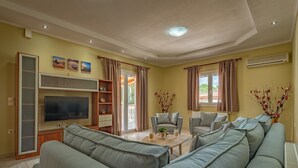 Una sala de estar con un sofá grande de color gris, una mesa de centro y dos sillones. Hay un televisor montado en la pared, una estantería y una ventana con cortinas.